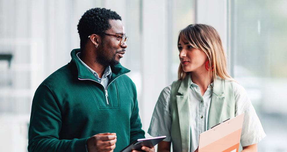 Two recent graduates in a bright office setting, talking while holding a tablet and documents.