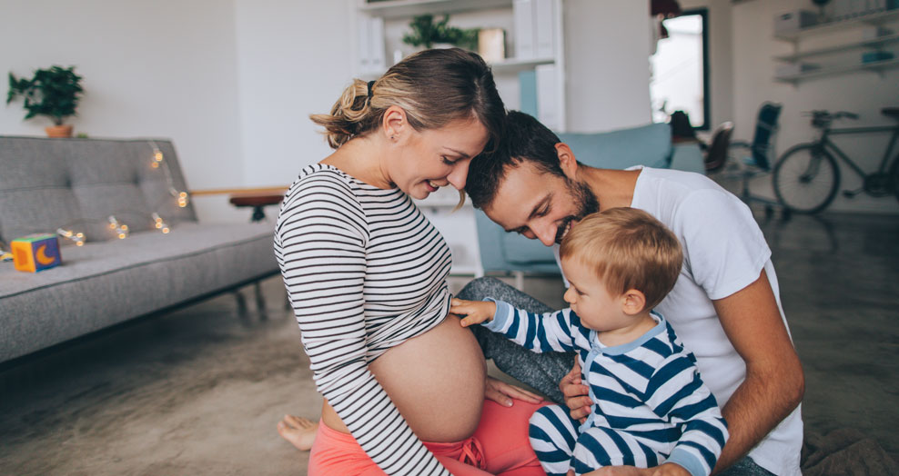 A child touches the belly of a pregnant person while another adult smiles beside them.